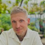 Ein lächelnder Mann mit weißen Haaren, einem weißen Hemd, Tattoos am Hals und einem Ohring.