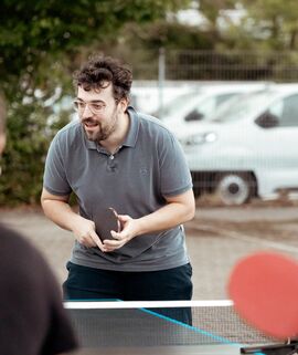 Männlich gelesene tischtennisspielende Person bei einer Firmenfeier für Mitarbeitende. Vier Personen spielen beim Herbstfest des Werksarztzentrums Deutschland Tischtennis an einer Tischtennisplatte.