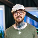 Ein sympathischer Mann mit einem Tattoo am Hals, einer silbernen Kette, einer braunen Brille, einer Mütze und einem grünen Shirt.