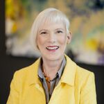 Ein Portraitfoto einer Frau in einem gelben Blazer und einer grauen Bluse. Sie lacht freundlich in die Kamera. Im Hintergrund befindet sich ein buntes Gemälde.