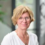 Eine freundlich lächelnde Frau mit blonden Haaren, weißer Bluse und Brille.