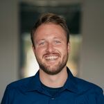 Ein Foto von Jonas Treixler. Er trägt ein blaues Hemd, auf dem seine Firma "SKOLAWORK" steht. Er grinst breit und freundlich direkt in die Kamera.