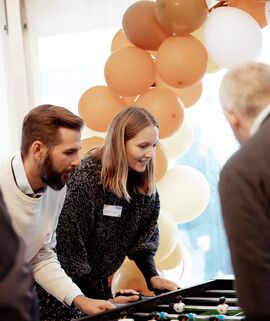 Vier Mitarbeitende kickern an einem Kickertisch. Im Hintergrund sind orangene Luftballons und Herbstdeko.