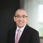 Foto eines lächelnden Mannes mit Brille. Er trägt einen schwarzen Anzug mit einem weißen Hemd und einer rosafarbenen Krawatte.