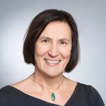 Eine lächelnde Frau mit einem dunkelbraunen Bob-Haarschnitt und einer ovalen, grünen Kette um den Hals.