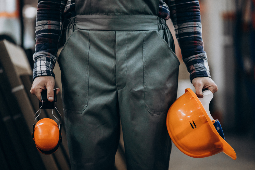 Person in einer Arbeitslatzhose mit einem orangenen Schutzhelm und Gehörschutz steht in einem Handwerksbetrieb.
