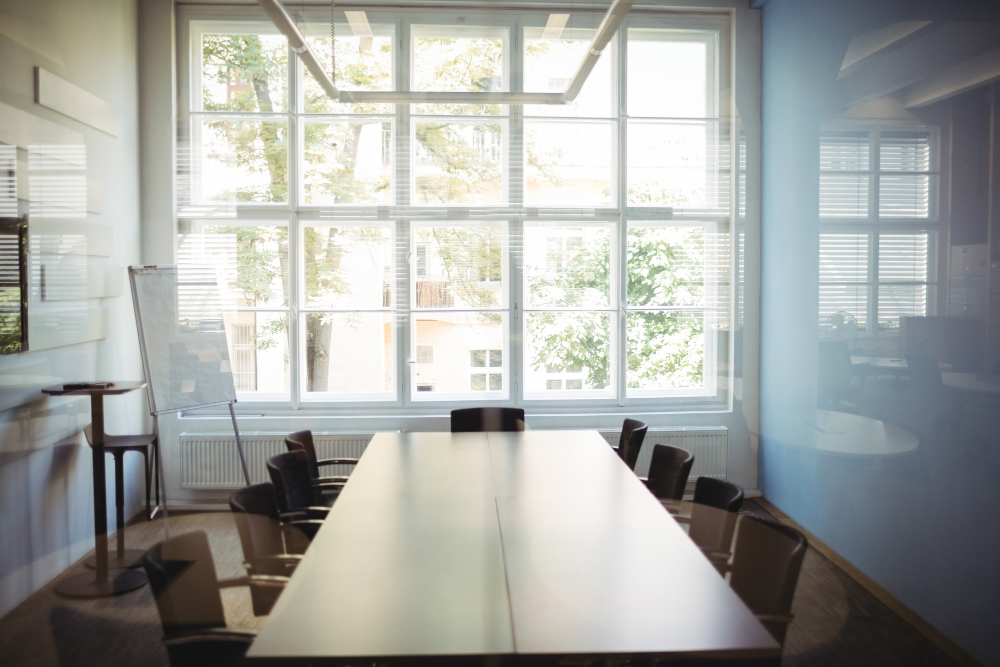 Leerer Meetingraum mit großer Glasfassade und vielen leeren Stühlen an einem länglichen Holztisch. Durch eine Scheibe fotografiert.