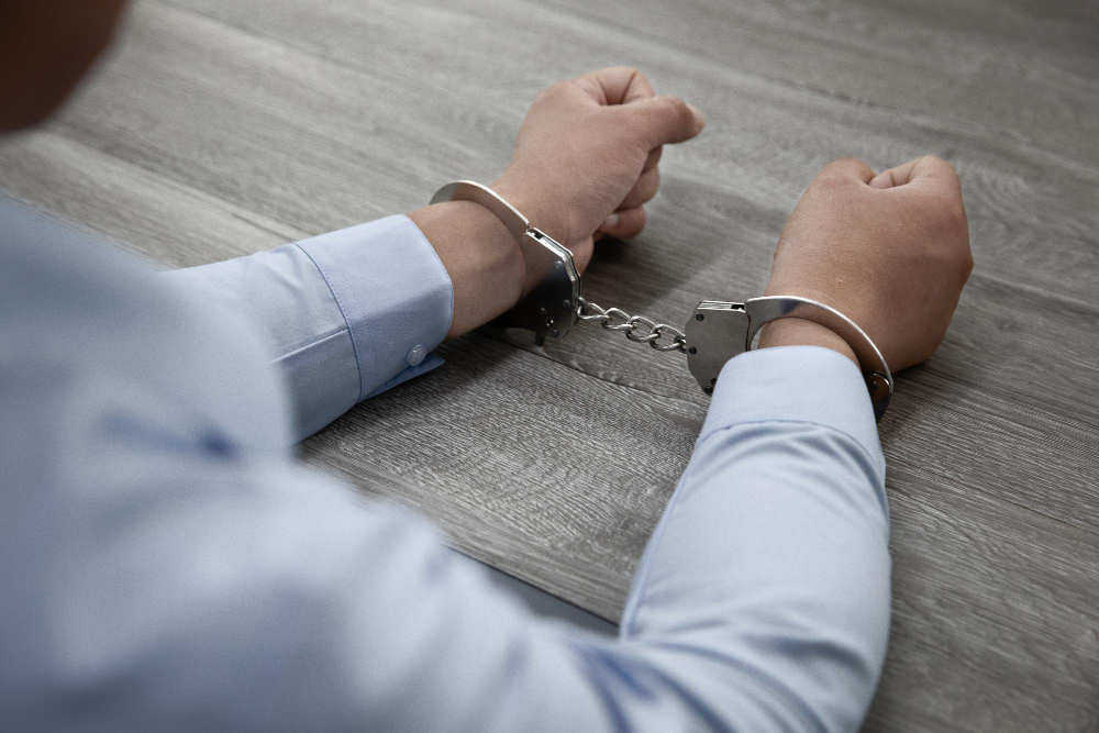 Ein Mensch in hellblauem Hemd, seine Hände sind in Handschellen und liegen vor ihm auf einem grauen Tisch.