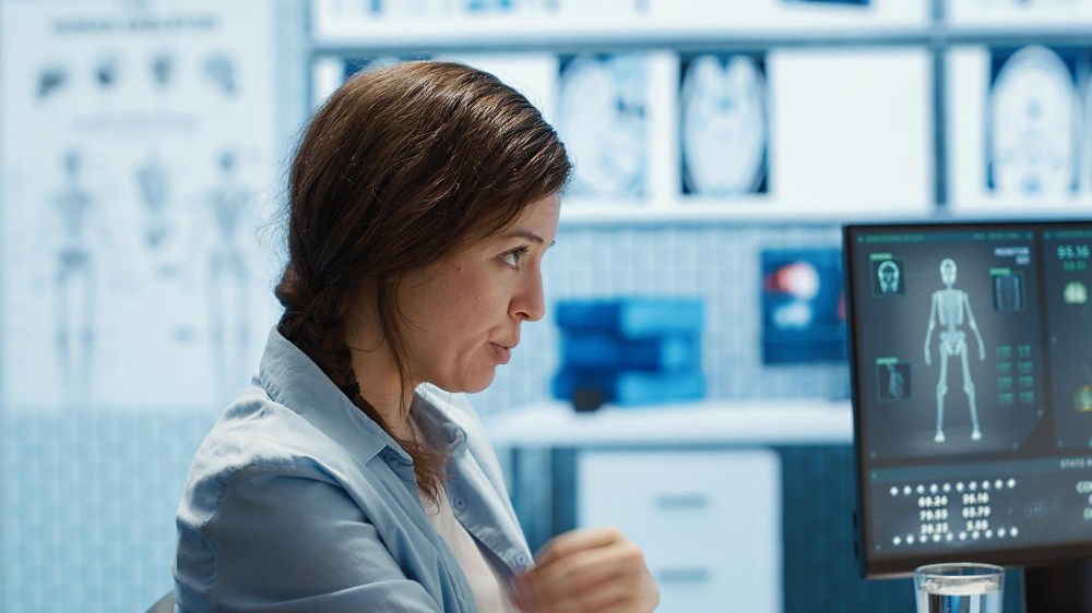 Eine Frau in einem Labor, rechts im Bild ein Bildschirm mit medizinischen Bildern, die von künstlicher Intelligenz generiert sind.