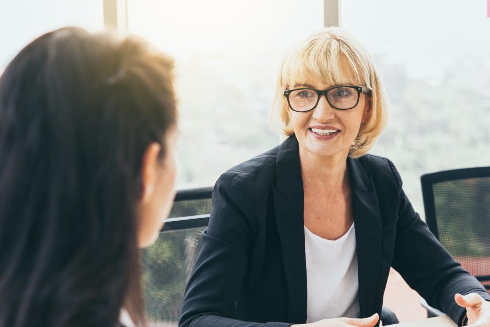 Zwei Frauen im Gespräch in einem Meetingraum. Die eine trägt eine Brille und einen schwarzen Blazer.