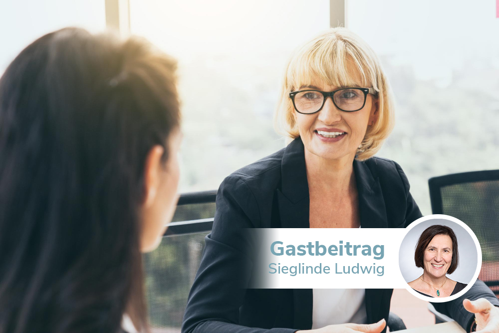 Zwei Frauen im Gespräch in einem Meetingraum. Die eine trägt eine Brille und einen schwarzen Blazer.