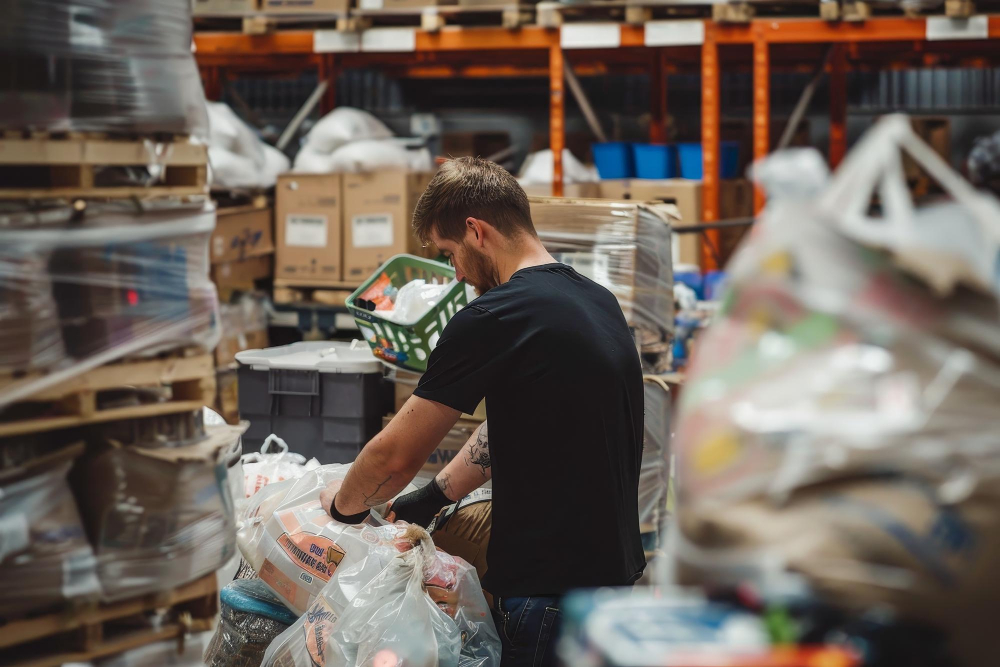 Ein Mann steht in einem Lager. Um ihn rum viele Paletten. Das Lager ist vollgestellt und chaotisch.