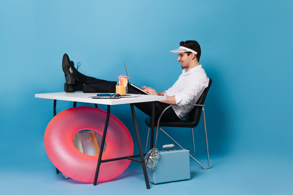 Mann sitzt mit einer Schirmmütze an in einem Studiokontext an einem Schreibtisch. Seine Füse hat er auf den Tisch gelegt und arbeitet mit einem Laptop auf den Beinen. Unter dem Tisch liegt rein pinker Schwimmring, ein Arbeitskoffer und eine Taucherbrille.