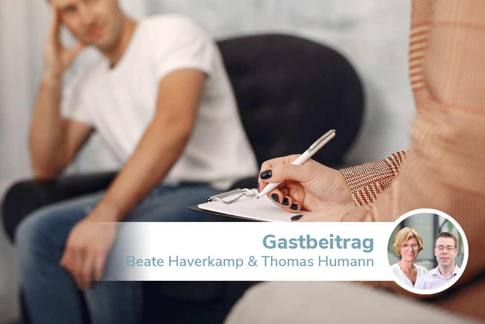 Eine Frau und ein Mann sitzen sich gegenüber. Die Frau notiert sich mit einem Kugelschreiber etwas auf einem Zettel auf einem Klammbrett. Der Mann im Hintergrund wirkt niedergeschlagen und nachdenklich.