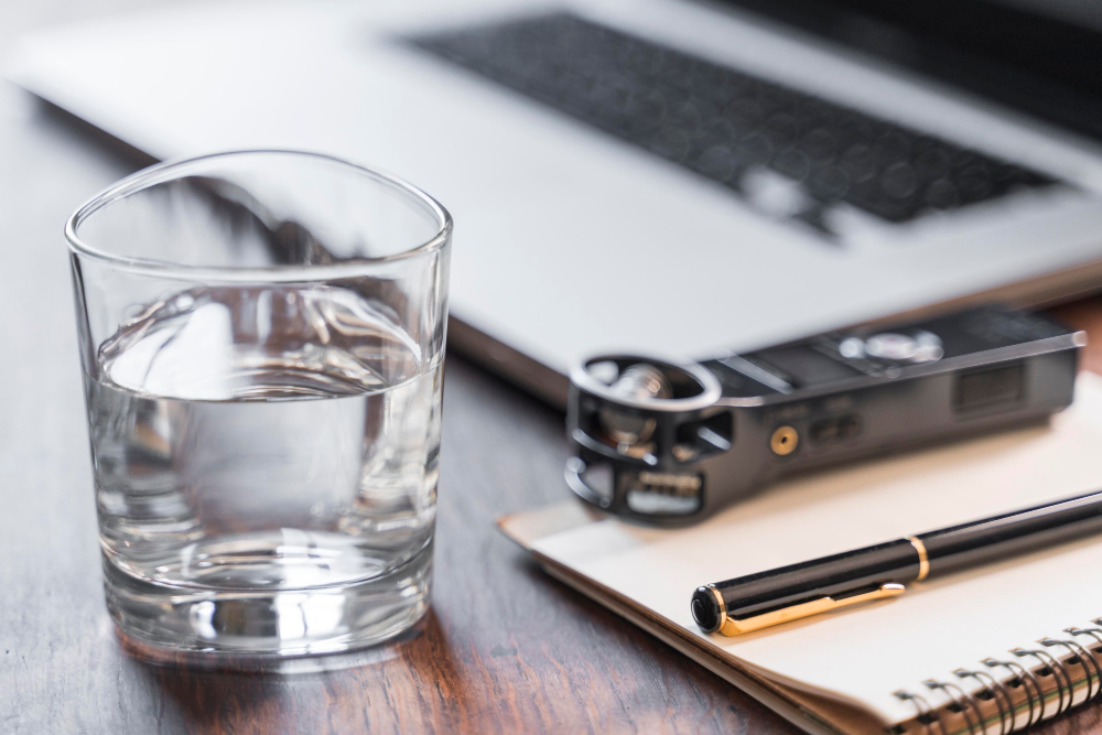 Ein Glas mit einer klaren Flüsigkeit steht auf einem Schreibtisch neben einem Laptop, einem Notizblock und einem schwarzen Kugelschreiben. Der Fokus des Fotos liegt auf dem Glas.