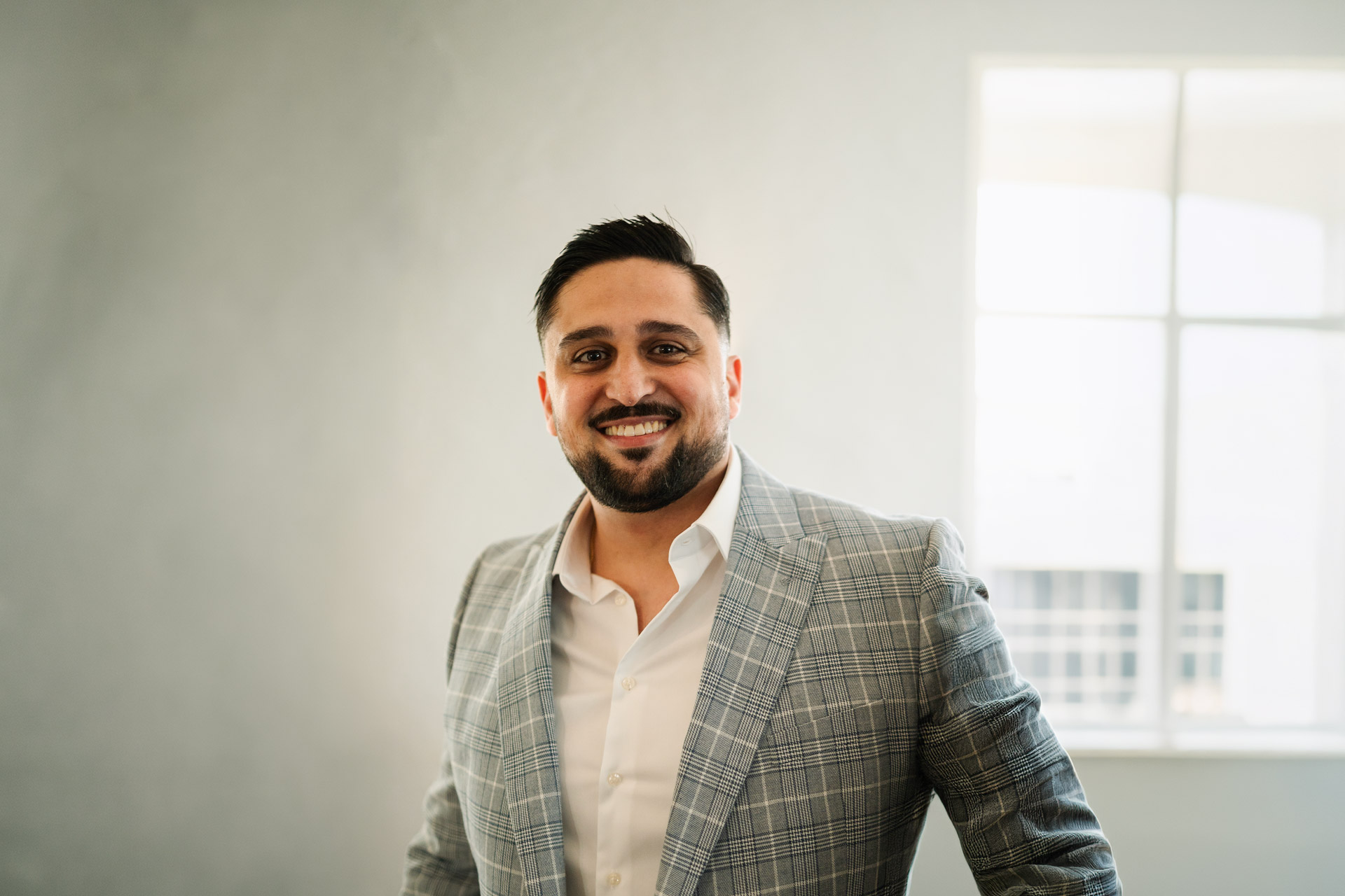 Arasch trägt ein kariertes Sakko zu einem weißen Hemd, er lacht herzerwärmend.