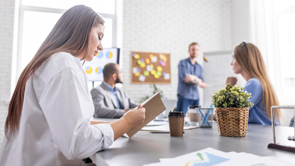 Eine Frau in weißer Bluse sitzt in einem Büro und schaut in ein Notizheft. Im Hintergrund reden drei weitere Personen vor einer Pinnwand mit bunten Post-Its.