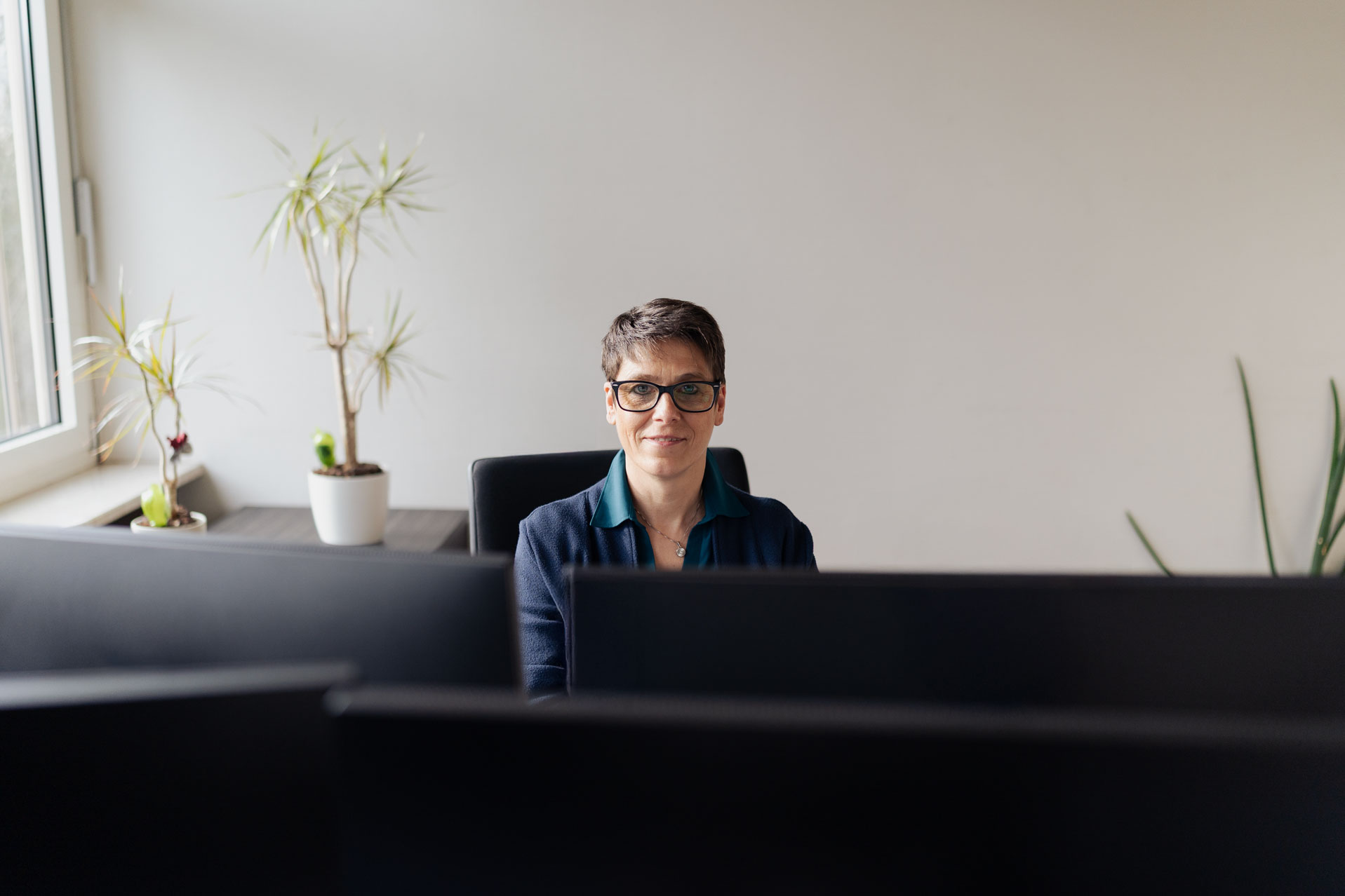 Stefanue sitzt an ihrem Arbeitsplatz. Links sehen wir eine Fensterbank mit Pflanzen. Sie selbst ist in der Mitte des Fotos mit einem blauen Oberteil und einer Brille.