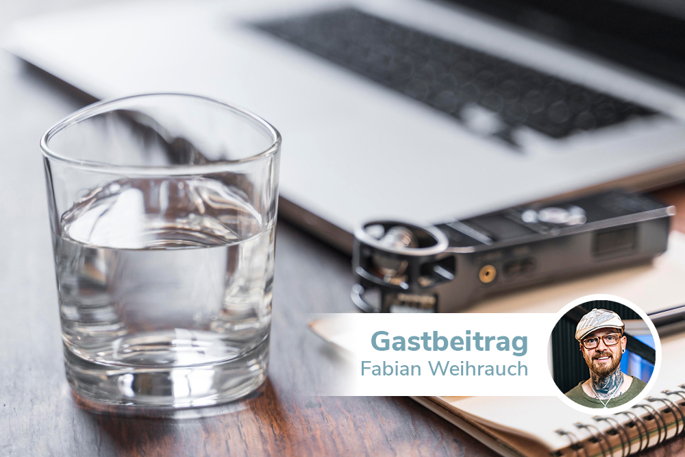 Ein Glas mit einer klaren Flüsigkeit steht auf einem Schreibtisch neben einem Laptop, einem Notizblock und einem schwarzen Kugelschreiben. Der Fokus des Fotos liegt auf dem Glas.