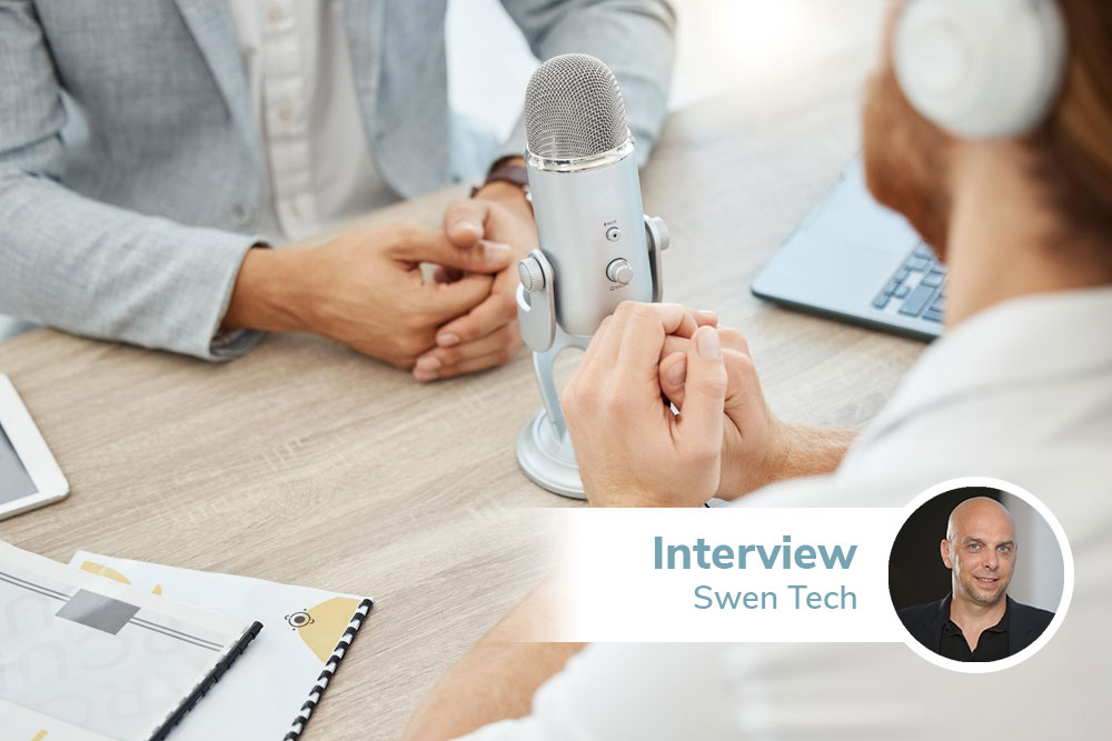 Ein silbernes Standmikrophon steht auf einem Tisch, auf beiden Seiten des Mirkophons sitzt eine Person.