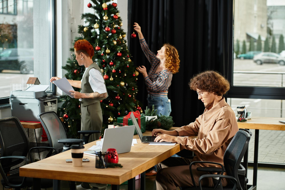 Ein Foto eines Büroraums, indem ein Mann an einem Laptop arbeitet, eine Frau etwas aus dem Drucker holt und eine andere Frau im Hintergrund eine Christbaumkugel an den Weihanchtsbaum im Bürogebäude hängt.