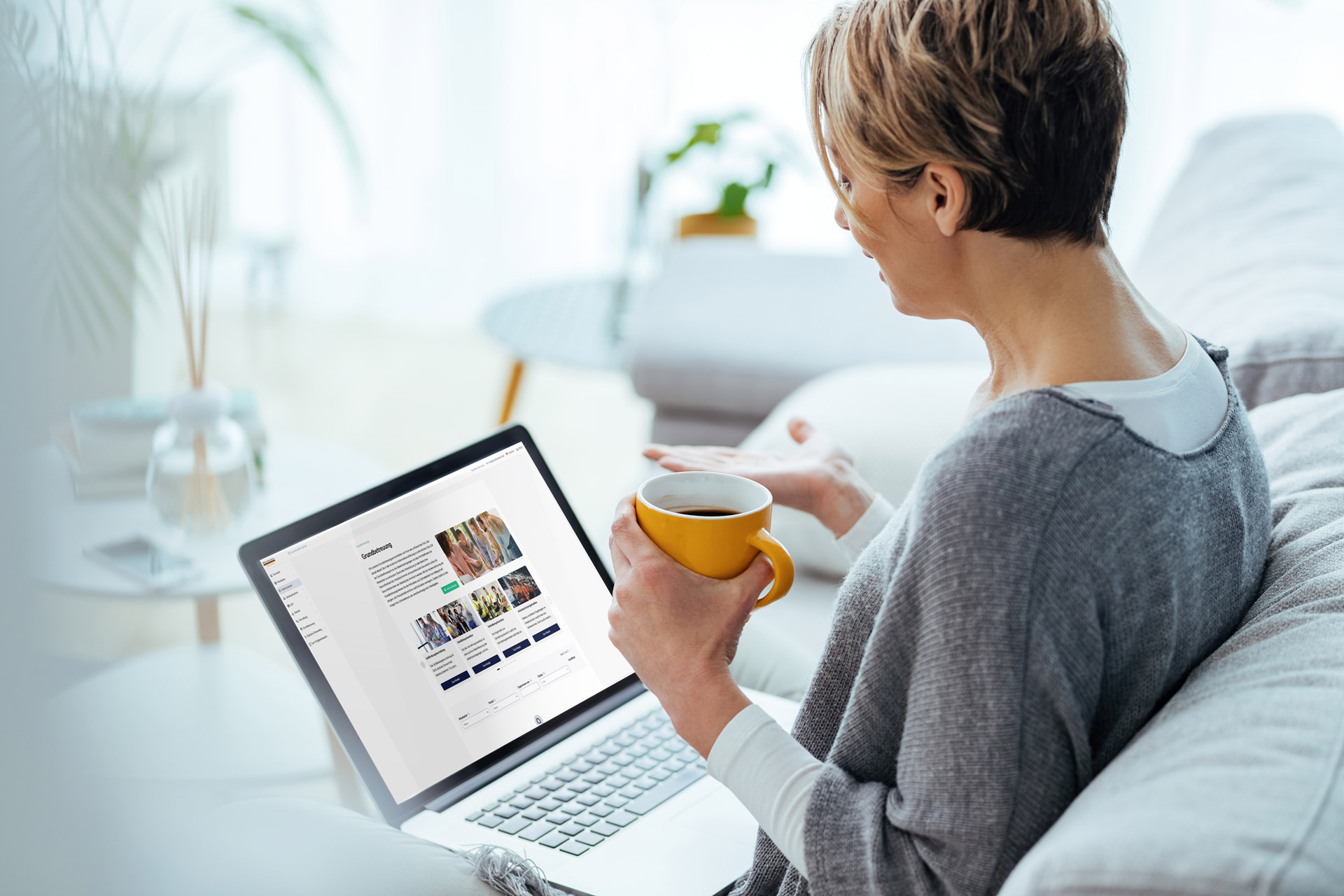 Eine Frau sitzt erkältet mit einer Tasse Tee auf dem Sofa zuhause und guckt sich auf dem Laptop das Portal des Werksarztzentrums Deutschland an.